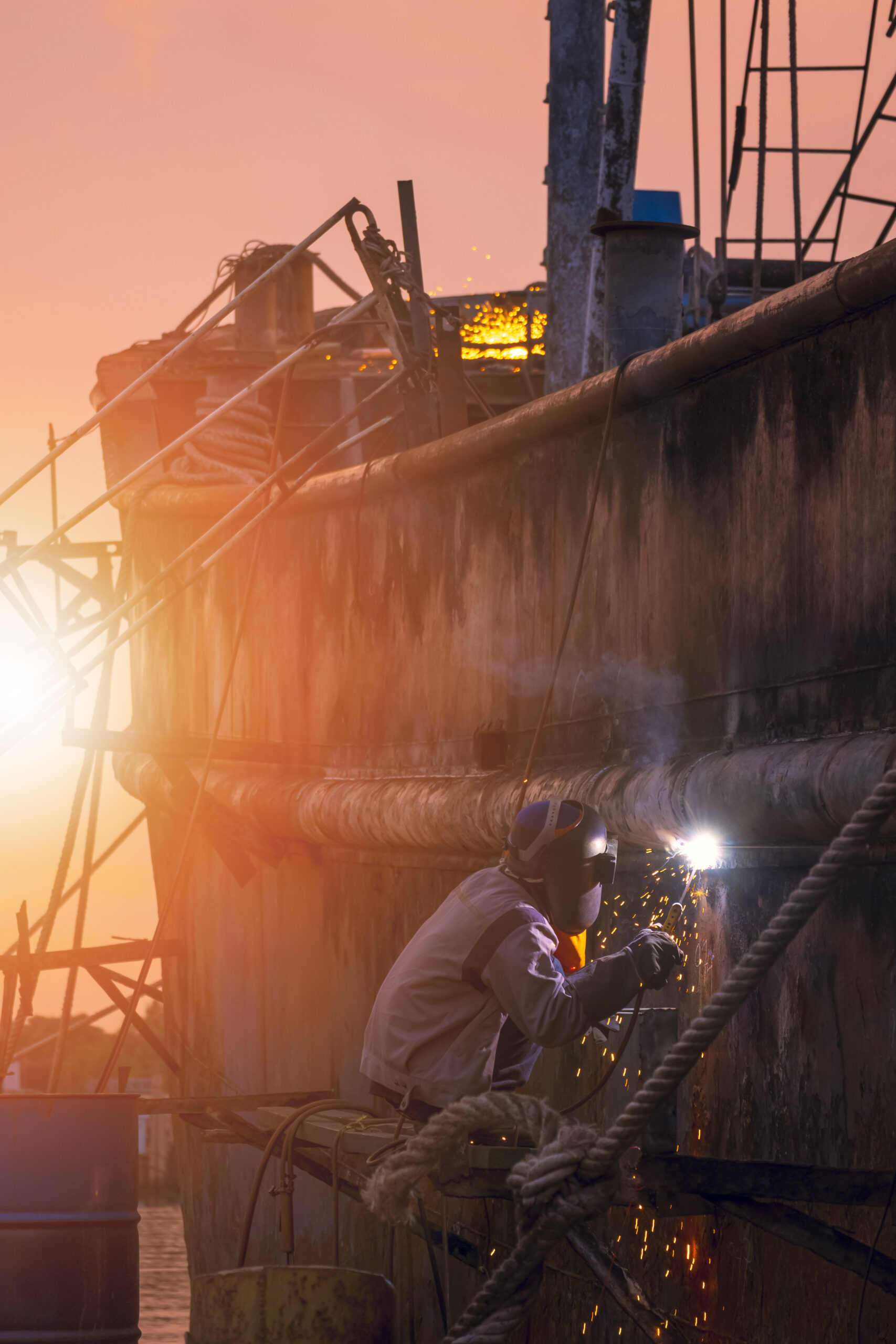 Side view of welder in protective workwear on platform is welding metal wall of the old fishing vessel in shipyard area at sunset time in vertical frame